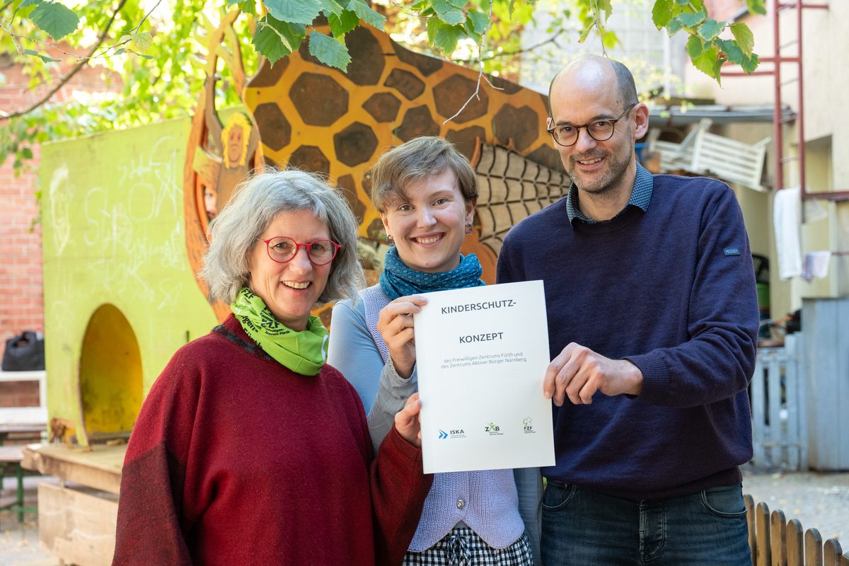 Die Leiterin des Freiwilligen-Zentrums Fürth und die beiden Projektverantwortlichen des Projekts Große für Kleine, das vom Engagement für und mit Kindern geprägt ist, stehen im Hof einer Kita und halten stolz das Kinderschutzkonzept hoch.