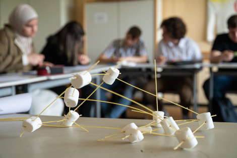 Im Vordergrund steht eine abstrakte Steck-Konstruktion aus Spaghetti und Marschmellows, im Hintergrund sitzen unscharf abgebildet Schüler/innen, die an etwas basteln.