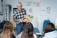 Ein langjähriger Ehrenamtlicher im Kita-Bereich steht in einem Klassenzimmer mit einer Schalttafel vor drei Mädchen und lächelt begeistert.