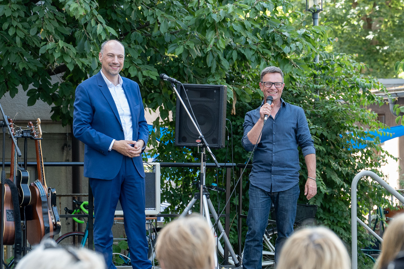 Zu sehen sind der zweite Bürgermeister, ein Mann mittleren Alters mit Halbglatze, und der Leiter des ZAB, ein Mann mittleren Alters mit Brille und blonden kurzen Haaren, auf einer Bühne im Freien während der Begrüßung. Beide tragen einen blauen Anzug und lächeln ins Publikum.
